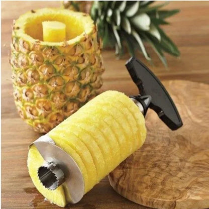Stainless steel corer holding a spiral-cut pineapple cylinder beside the hollowed fruit on a wooden board